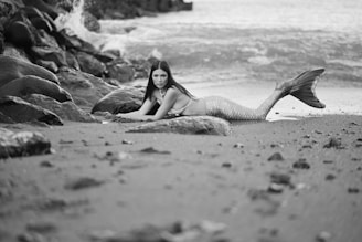 A mermaid with long dark hair lies on a rocky beach near the water's edge, surrounded by scattered rocks. The scene is captured in black and white, giving it a classic and timeless feel.
