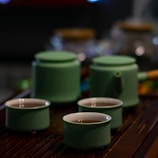 A tea set consisting of three green ceramic cups filled with tea and a matching teapot with a wooden handle. The set is placed on a dark wooden surface with out-of-focus glass jars in the background, creating a cozy atmosphere.