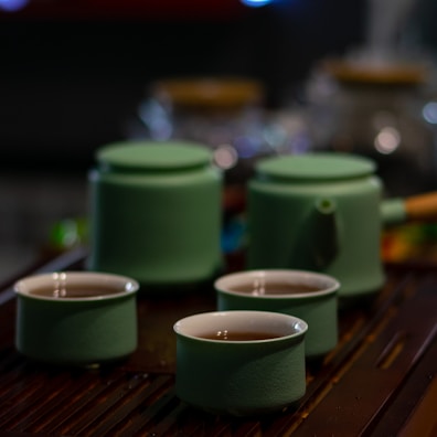 A tea set consisting of three green ceramic cups filled with tea and a matching teapot with a wooden handle. The set is placed on a dark wooden surface with out-of-focus glass jars in the background, creating a cozy atmosphere.