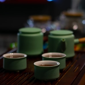 A tea set consisting of three green ceramic cups filled with tea and a matching teapot with a wooden handle. The set is placed on a dark wooden surface with out-of-focus glass jars in the background, creating a cozy atmosphere.