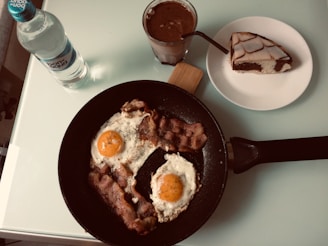 A breakfast setting featuring a non-stick pan with two sunny-side-up eggs and crispy bacon garnished with herbs. Nearby is a bottle of mineral water and a glass of chocolate drink. A slice of marble cake is placed on a white plate.