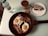 A breakfast setting featuring a non-stick pan with two sunny-side-up eggs and crispy bacon garnished with herbs. Nearby is a bottle of mineral water and a glass of chocolate drink. A slice of marble cake is placed on a white plate.