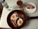A breakfast setting featuring a non-stick pan with two sunny-side-up eggs and crispy bacon garnished with herbs. Nearby is a bottle of mineral water and a glass of chocolate drink. A slice of marble cake is placed on a white plate.