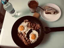 A breakfast setting featuring a non-stick pan with two sunny-side-up eggs and crispy bacon garnished with herbs. Nearby is a bottle of mineral water and a glass of chocolate drink. A slice of marble cake is placed on a white plate.