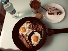 A breakfast setting featuring a non-stick pan with two sunny-side-up eggs and crispy bacon garnished with herbs. Nearby is a bottle of mineral water and a glass of chocolate drink. A slice of marble cake is placed on a white plate.