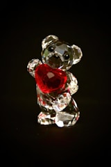 A small crystal bear figurine holds a bright red heart against a dark background. The bear is faceted, catching light in a way that creates a sparkling effect.