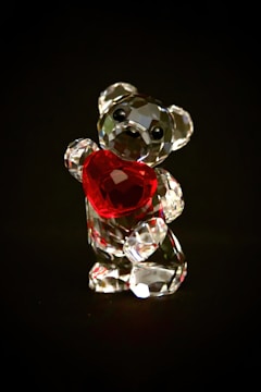 A small crystal bear figurine holds a bright red heart against a dark background. The bear is faceted, catching light in a way that creates a sparkling effect.