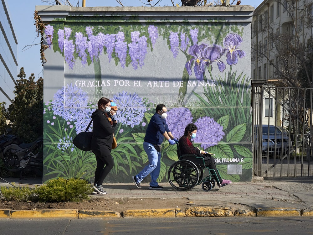 a group of people walking down a sidewalk next to a wall with flowers