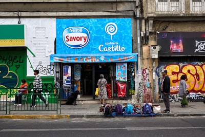 A small convenience store with a bright blue sign above the entrance displays 'Nestlé Savory' and 'confitería Castillo.' People are walking by or standing outside on the sidewalk. The storefront is surrounded by colorful graffiti and urban art. There are several backpacks and bags laid out on the pavement in front of the store.