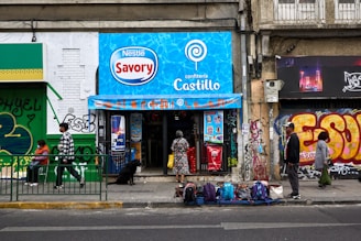 A small convenience store with a bright blue sign above the entrance displays 'Nestlé Savory' and 'confitería Castillo.' People are walking by or standing outside on the sidewalk. The storefront is surrounded by colorful graffiti and urban art. There are several backpacks and bags laid out on the pavement in front of the store.