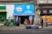 A small convenience store with a bright blue sign above the entrance displays 'Nestlé Savory' and 'confitería Castillo.' People are walking by or standing outside on the sidewalk. The storefront is surrounded by colorful graffiti and urban art. There are several backpacks and bags laid out on the pavement in front of the store.