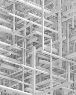 An intricate, abstract structure composed of interconnected, three-dimensional rods and beams. The arrangement creates a complex, geometric pattern with overlapping and intersecting lines, giving a sense of depth and density. The monochromatic color scheme emphasizes the shapes and shadows within the structure.