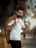 A man with a neatly trimmed beard and styled hair is holding sunglasses in one hand while draping a brown jacket over his shoulder. He wears a white t-shirt and black pants, standing in a blurred outdoor setting that suggests a calm and relaxed atmosphere.