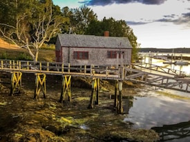 A rustic wooden cabin sits on stilts above a rocky shoreline. The structure is connected to the shore by a weathered wooden walkway. Surrounding the cabin are lush green trees and bushes. In the background, a body of water reflects the soft light of the setting or rising sun, and several small boats are docked along another pier.