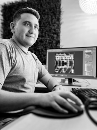 A person wearing a gray t-shirt is seated at a desk, using a mouse and keyboard in front of a computer monitor. The monitor displays graphic design software with text in a different language. The setting appears professional, with a blurred background possibly featuring office decor.