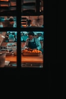 A chef is focused on preparing food in a dimly lit kitchen, surrounded by kitchen tools and ingredients. The scene is viewed through a window with a grid pattern.