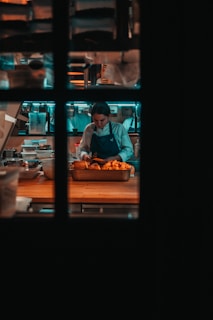 A chef is focused on preparing food in a dimly lit kitchen, surrounded by kitchen tools and ingredients. The scene is viewed through a window with a grid pattern.