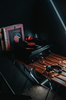 A vintage-style setup featuring a turntable playing a vinyl record sits on a wooden table. Next to the turntable are several vinyl record covers, one prominently displayed with a dark color scheme. A pair of headphones rests on the table, connected to the turntable. The scene is dimly lit, creating a cozy, nostalgic atmosphere.