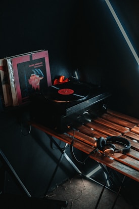A vintage-style setup featuring a turntable playing a vinyl record sits on a wooden table. Next to the turntable are several vinyl record covers, one prominently displayed with a dark color scheme. A pair of headphones rests on the table, connected to the turntable. The scene is dimly lit, creating a cozy, nostalgic atmosphere.