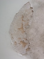 A section of wall with peeling paint, revealing the underlying plaster. The texture is rough and uneven, with patches of discoloration.