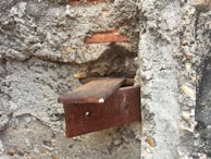 An old, rusted metal bracket is embedded in a textured concrete wall. The surface of the wall is rough, with visible pebbles and chunks of cement. The metal shows significant corrosion, adding a rustic and weathered appearance.