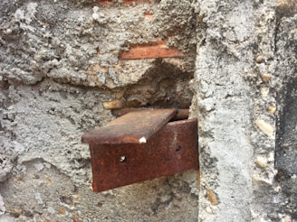 An old, rusted metal bracket is embedded in a textured concrete wall. The surface of the wall is rough, with visible pebbles and chunks of cement. The metal shows significant corrosion, adding a rustic and weathered appearance.