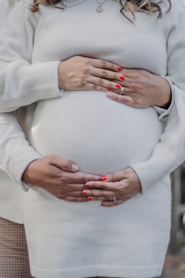 A close-up view of a pregnant person in a light-colored sweater, with a pair of hands with red painted nails gently resting on the belly, suggesting a connection or support. Another pair of hands is placed closely below, clasping the first pair.
