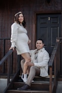 A couple is posing together on a wooden staircase in front of a dark door. The woman is pregnant, wearing a light-colored dress, and has one foot on a step while the man is kneeling next to her, wearing a light-colored jacket and brown pants. They both appear to be smiling.
