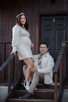 A couple is posing together on a wooden staircase in front of a dark door. The woman is pregnant, wearing a light-colored dress, and has one foot on a step while the man is kneeling next to her, wearing a light-colored jacket and brown pants. They both appear to be smiling.