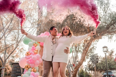 A joyful couple is celebrating outdoors, holding smoke bombs emitting vibrant pink and purple clouds. They are standing in front of a colorful balloon arrangement with shades of pink, green, and gold. Trees and a clear sky are visible in the background, creating a festive atmosphere.