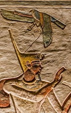 An ancient Egyptian wall carving depicts a figure wearing a tall, ornate headdress, possibly a pharaoh or deity. A bird with outstretched wings is also carved, appearing above the figure's head. The carvings are detailed with distinct lines and colors, highlighting traditional Egyptian art style.