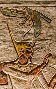 An ancient Egyptian wall carving depicts a figure wearing a tall, ornate headdress, possibly a pharaoh or deity. A bird with outstretched wings is also carved, appearing above the figure's head. The carvings are detailed with distinct lines and colors, highlighting traditional Egyptian art style.