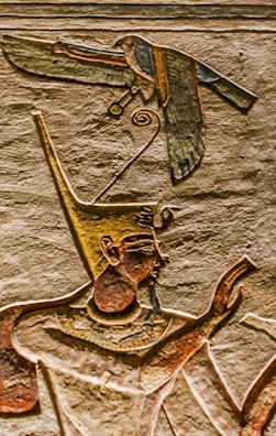 An ancient Egyptian wall carving depicts a figure wearing a tall, ornate headdress, possibly a pharaoh or deity. A bird with outstretched wings is also carved, appearing above the figure's head. The carvings are detailed with distinct lines and colors, highlighting traditional Egyptian art style.