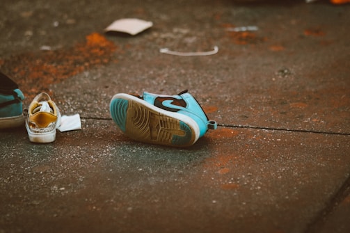 A pair of sneakers is lying on a rough, textured concrete surface. One shoe is upside down revealing its sole, which is beige in color. The other shoe is upright and features a colorful design with blue and yellow elements. The ground is speckled with orange and white markings, adding a gritty feel to the scene.