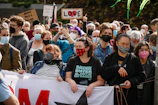 A diverse crowd of people, some wearing masks, gathered outdoors for a protest or rally. The individuals hold a large banner and signs with various messages. The mood appears energetic and positive as people express their views.