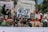 A gathering of people in an outdoor space, holding signs with messages such as 'Keep Your Roleplay in the Bedroom' and wearing masks. The scene appears to be a protest or demonstration, with multiple individuals engaged in conversations and standing in groups. There are banners and flags visible in the background.
