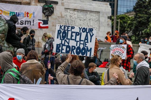 A gathering of people in an outdoor space, holding signs with messages such as 'Keep Your Roleplay in the Bedroom' and wearing masks. The scene appears to be a protest or demonstration, with multiple individuals engaged in conversations and standing in groups. There are banners and flags visible in the background.