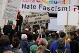A crowd of people are gathered at a protest holding various signs with slogans such as 'Freedom of Religion Equals Freedom From Religion' and 'For massive wage and benefit increases!'. A large banner in the background states messages against fascism and in support of community.