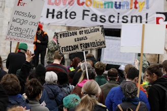 A crowd of people are gathered at a protest holding various signs with slogans such as 'Freedom of Religion Equals Freedom From Religion' and 'For massive wage and benefit increases!'. A large banner in the background states messages against fascism and in support of community.