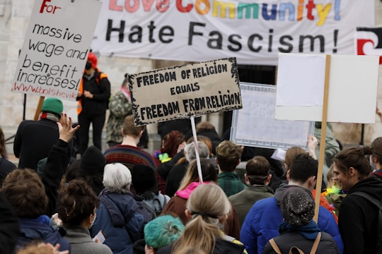 A crowd of people are gathered at a protest holding various signs with slogans such as 'Freedom of Religion Equals Freedom From Religion' and 'For massive wage and benefit increases!'. A large banner in the background states messages against fascism and in support of community.