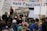 A crowd of people are gathered at a protest holding various signs with slogans such as 'Freedom of Religion Equals Freedom From Religion' and 'For massive wage and benefit increases!'. A large banner in the background states messages against fascism and in support of community.