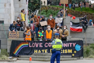 A group of people are gathered on a set of stone steps, holding various signs and banners with messages such as 'LOVE COMMUNITY HATE RACISM', 'UNITE AGAINST FASCISM', and 'WHO ELECTED YOU?'. Most people are wearing masks and some are wearing colorful clothing. A police officer stands in the foreground, observing the crowd.