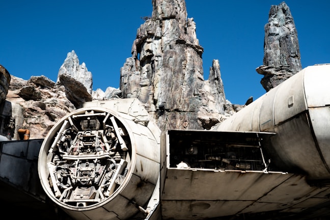 A detailed view of a spacecraft with intricate machinery exposed in the foreground. Towering rocky formations in the background under a clear blue sky create a dramatic scene.