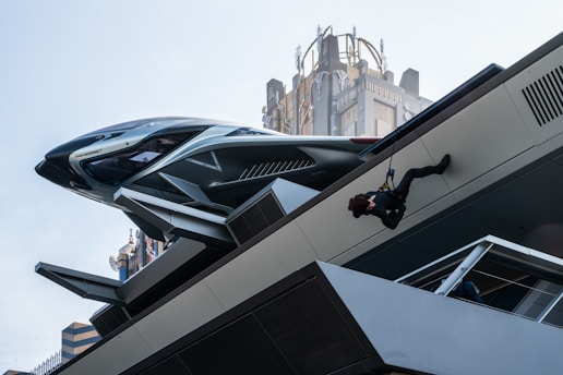A futuristic setting with a sleek, advanced vehicle or spacecraft parked on a platform. A person is suspended in mid-air, climbing or rappelling down a structure. The background features a tall building with intricate architectural details.
