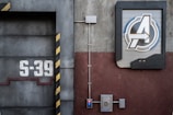 A detailed view of an industrial-style wall with a large emblem featuring a stylized letter 'A' within a circular frame, positioned on the right side. To the left, the number 'S-39' is prominently displayed on a metallic surface with black and yellow hazard stripes. Electrical panels and conduits run along the surface, contributing to the mechanical and utilitarian aesthetic.