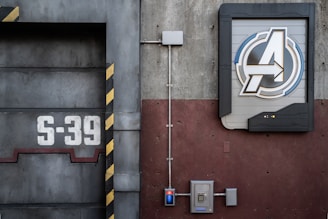 A detailed view of an industrial-style wall with a large emblem featuring a stylized letter 'A' within a circular frame, positioned on the right side. To the left, the number 'S-39' is prominently displayed on a metallic surface with black and yellow hazard stripes. Electrical panels and conduits run along the surface, contributing to the mechanical and utilitarian aesthetic.