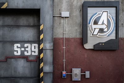 A detailed view of an industrial-style wall with a large emblem featuring a stylized letter 'A' within a circular frame, positioned on the right side. To the left, the number 'S-39' is prominently displayed on a metallic surface with black and yellow hazard stripes. Electrical panels and conduits run along the surface, contributing to the mechanical and utilitarian aesthetic.