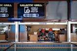 Two workers in orange caps and uniforms are preparing and handing over food at a counter labeled 'Galactic Grill' with signage indicating mobile order pick-up at window 5. The scene is set in a food service area with multiple signs, trays of food, and various utensils on the countertop.