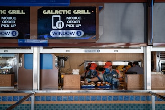 Two workers in orange caps and uniforms are preparing and handing over food at a counter labeled 'Galactic Grill' with signage indicating mobile order pick-up at window 5. The scene is set in a food service area with multiple signs, trays of food, and various utensils on the countertop.