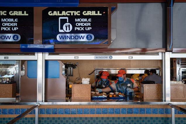 Two workers in orange caps and uniforms are preparing and handing over food at a counter labeled 'Galactic Grill' with signage indicating mobile order pick-up at window 5. The scene is set in a food service area with multiple signs, trays of food, and various utensils on the countertop.
